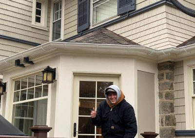 A smiling figure giving a thumbs-up outside of a beige house. The house has black shutters and white gutters